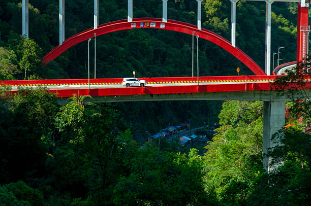 Beautiful mountain scenery with a striking red bridge known as Kelok 9 (Kelok Sembilan) in West Sumatra, Indonesia. The bridge winds gracefully through lush green hills and stretches over a deep valley with a river flowing below.