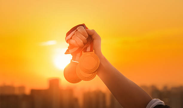 Raised hand holding three gold medal against sunset sky
