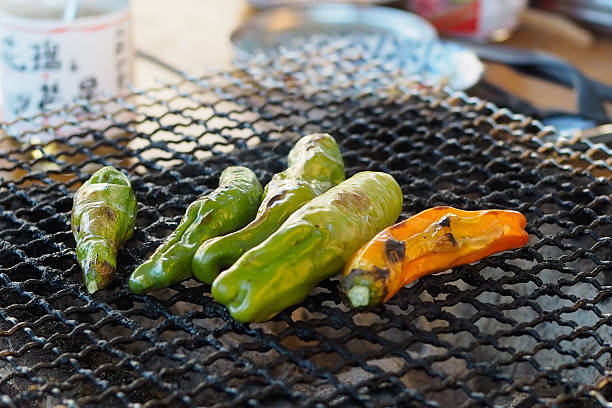 Fresh shishito peppers grilling over a traditional Japanese charcoal mesh. A simple, rustic BBQ scene with blistered skins and savory aroma in an outdoor setting.