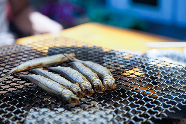 Whole shishamo fish being grilled over a traditional charcoal mesh grill. A rustic Japanese-style outdoor BBQ setting with natural lighting and authentic cooking.