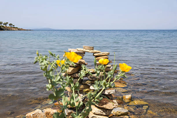 Close-up view of a small arch built from stacked flat stones, placed near the seashore with calm water in the background. Bright yellow wildflowers bloom in front, adding a touch of color to the natural setting. A peaceful and harmonious scene symbolizing balance, nature, and mindfulness.