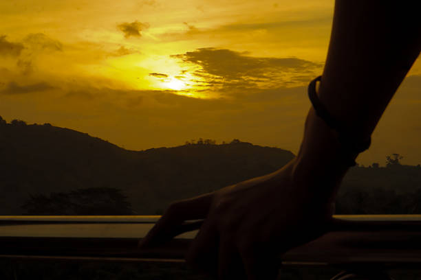 A dramatic silhouette of a hand gripping an iron fence, facing toward a hill during golden sunset. The image evokes emotion, solitude, and contemplation, with warm tones and scenic beauty.