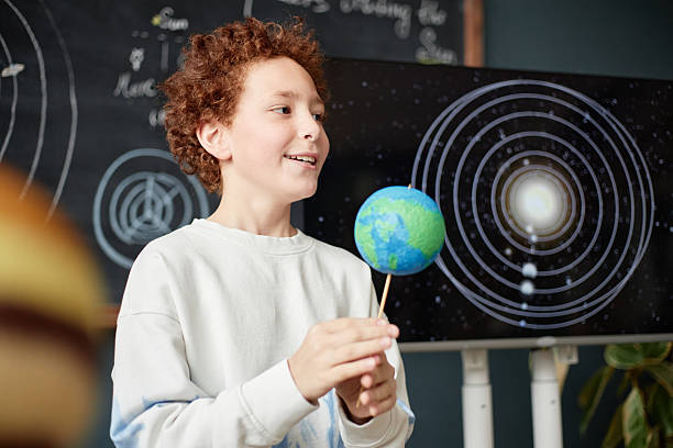 Caucasian boy holding model of Earth while standing in front of solar system diagram on screen, smiling and engaging in science activity, demonstrating interest in astronomy and education