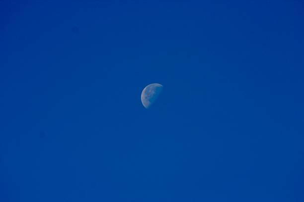 The moon is visible in a clear blue sky during daytime, showcasing a partial phase. The serene view captures the quiet beauty of the moon contrasting against a deep, cloudless sky.
