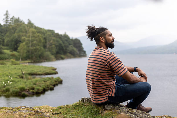 A wide shot of a man sitting down on a rocky surface at the waters edge of Derwent Water in Keswick, Cumbria. He is wearing a striped t-shirt and hiking boots as he is on a hike. The man is taking a moment of reflection as he looks out at the water. Trees and mountain ranges can be seen in the background under an overcast sky.Videos are available similar to this scenario.