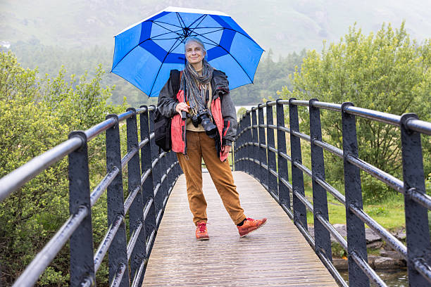 A wide portrait shot of a female wildlife photographer standing on a bridge in the countryside in Keswick, Cumbria. She is standing in casual clothing with a waterproof coat while holding a blue umbrella. The woman has a camera around her neck and is looking directly at the camera while smiling.Videos are available similar to this scenario.