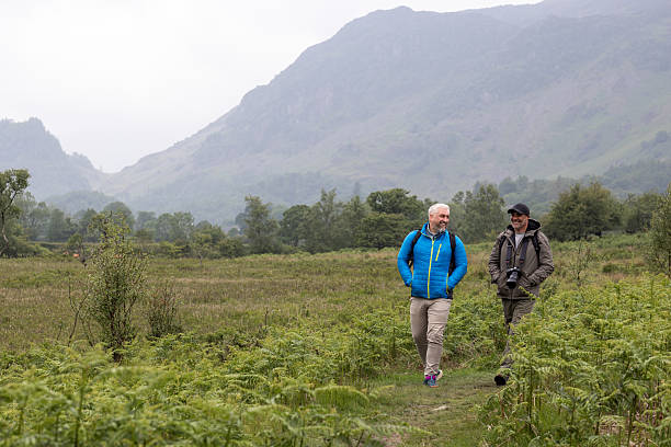 A wide shot of two men walking and talking on a grass path through a rural landscape in Keswick with tall grass and mountains in the background. They are wearing casual outdoor hiking clothing and backpacks, enjoying a peaceful hike in nature. The scene conveys male friendship, adventure, and healthy living.Videos are available similar to this scenario.
