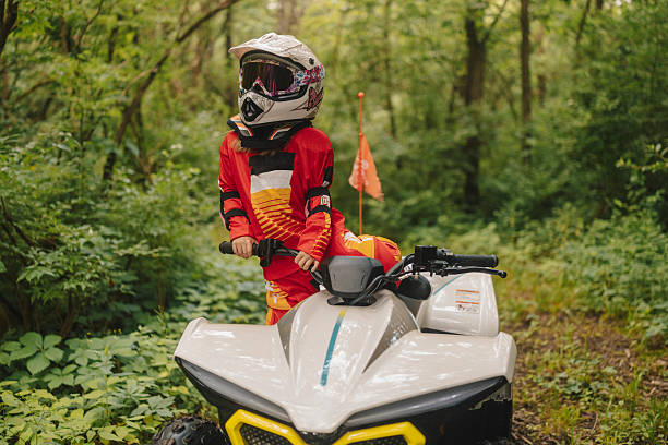 Little girl riding a child-sized ATV through a lush green forest during a camping trip. Wearing full protective gear including a helmet, she enjoys an exciting outdoor adventure surrounded by nature