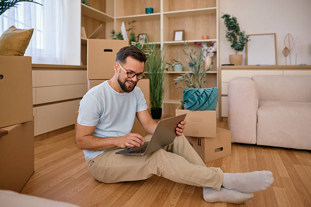 istock Man sitting on floor using laptop surrounded by cardboard boxes in new home 2225554185