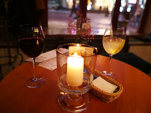 istock Burning thick candle inside transparent candle jar (photophore) framed by two glasses - with red wine and white cocktail as well some table ware in blurry background of big window with evening street and passing people in Moscow. 2225556513