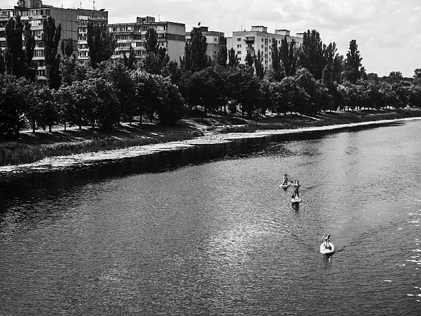 Black and white image of athletes paddling on their stand up paddle boards on a river near a residential area with apartment buildings