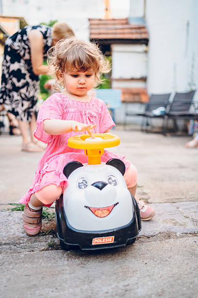 A cheerful little baby girl is playing outdoor. She driving little car in garden.