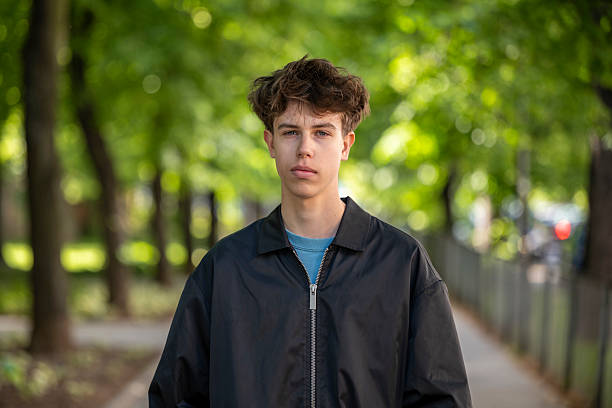 Portrait of a teenage boy standing confidently on a tree-lined city sidewalk. Natural light and casual expression reflect youth and street style.