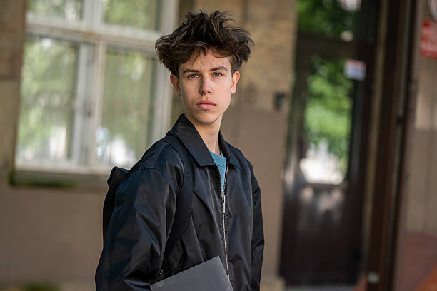Young man in black jacket looks directly at camera with a calm, serious expression. Urban background, natural light.