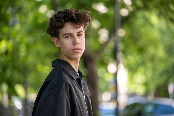 Pensive teenage boy with tousled hair and documents under his arm standing on a sunny park pathway.
