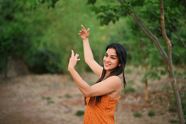 A cheerful young woman dances gracefully in a lush green forest. Her expressive hands and smiling face show she’s enjoying the peaceful, happy moment outdoors.