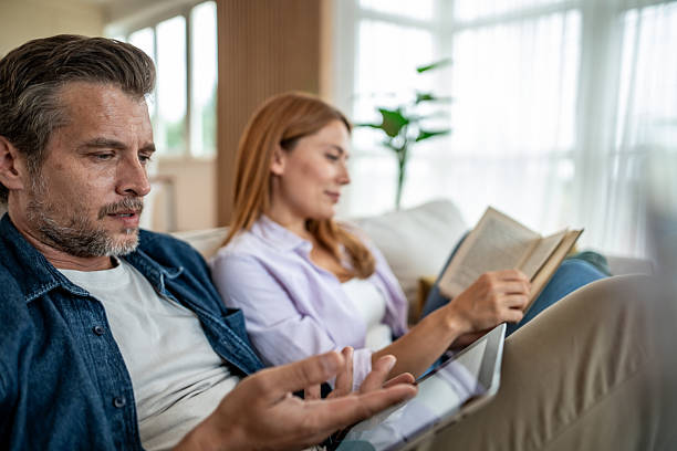 istock Couple relaxing on sofa: man using digital tablet, woman reading book 2225572742