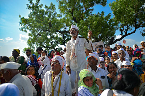An elderly man dressed in traditional white attire and Gandhi cap captures a moment on his smartphone while standing among a vibrant crowd of Varkari pilgrims during the Pandharpur Wari pilgrimage in Maharashtra, India. The image highlights the intersection of age-old spiritual traditions and modern technology, set against the backdrop of a lively outdoor gathering beneath a lush green tree.