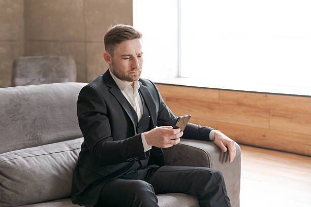 istock A man in a formal suit is sitting on a sofa in a bright office, staring intently at a mobile phone, reflecting business activity and confidence 2225580705