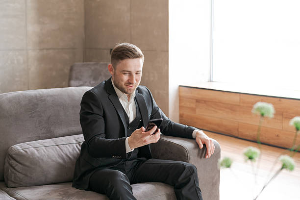 istock A man in a black suit is sitting on a couch using a smartphone in a modern interior with a large window 2225580715