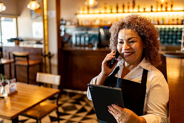 istock Senior waitress using digital tablet and talking on mobile phone at coffee shop 2225583420