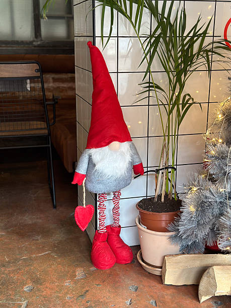 Christmas gnome with a bright red hat and white beard is positioned next to a potted palm. Festive ambiance warm lighting and a cozy interior