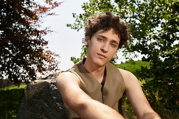 Young man with curly hair relaxes outdoors, showcasing his stylish summer outfit in the park.