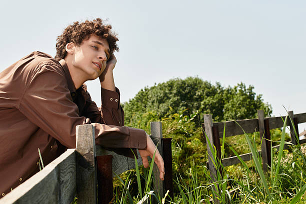 Curly-haired young man relaxes by a wooden fence on a sunny day.