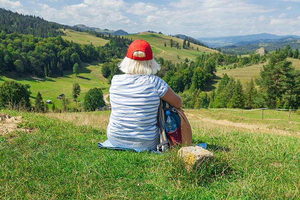 A senior woman with a red cap and striped shirt sits peacefully on a grassy hillside, gazing out at a stunning panoramic view of green valleys and distant mountain ranges. The photo captures a quiet moment of reflection during a nature hike, symbolizing freedom, mindfulness, and the joy of outdoor exploration in later life.