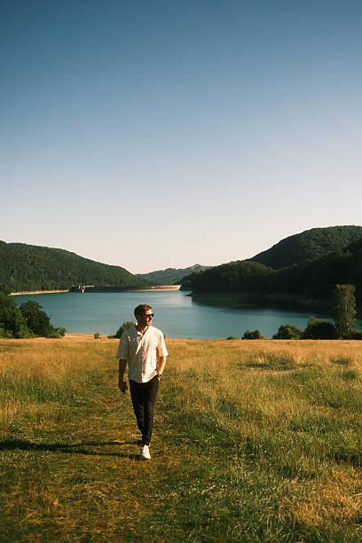 A man is leisurely walking in a beautiful field that features a serene lake situated in the picturesque background behind him