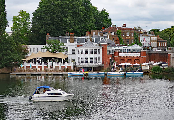 Hampton Court, London Bourgh of Richmond, United Kingdom, July 6, 2025. River Thames waterfront view from A309 road bridge. Boats, trees, clouds and riverside pubs. Cloudy summer day outdoors.