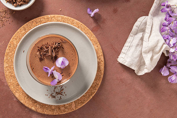 istock An overhead view features a single creamy chocolate mousse in a glass, styled on plates with a cork placemat, a bowl of shavings, and purple flowers. 2225627081