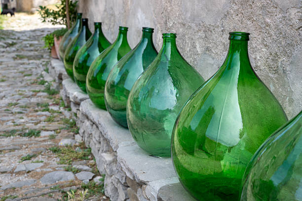 Some large green bulbous bottles stand on a low old wall on a house wall with part of an alley
