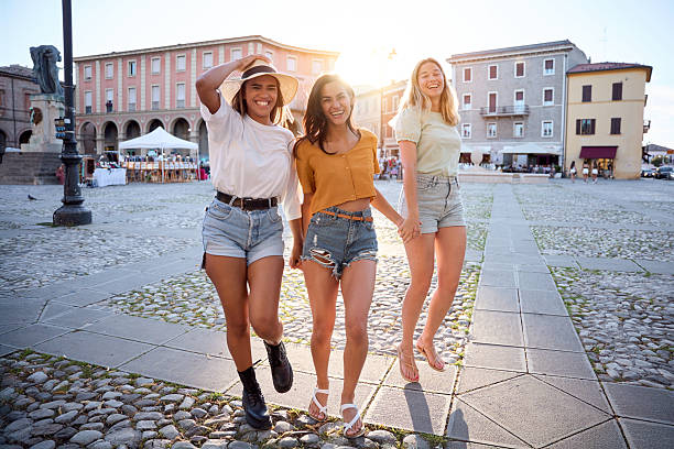 Three cheerful young women tourists are enjoying their vacation, holding hands and walking through a picturesque town square, bathed in the warm glow of the setting sun