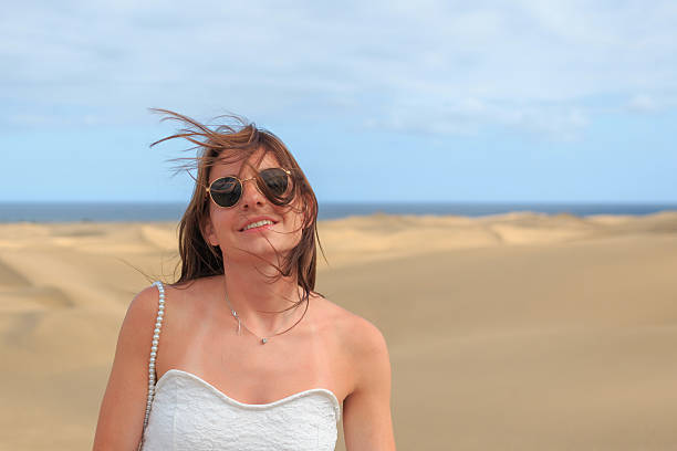 A confident woman in a white cropped top and wide-leg pants stands on a sunlit desert dune, shielding her eyes from the bright sun. The expansive blue sky highlights fitness, outdoor lifestyle, and wellness. Perfect for sports, travel, or inspirational content featuring strength and outdoor adventure.