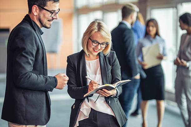 Caucasian business executives are engaging in a collaborative discussion at a conference. A woman is writing in a notebook while a man attentively listens. The setting is a professional networking event with blurred figures in the background.