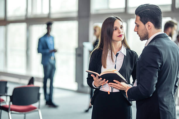 Caucasian business executives in formal attire discuss notes from a notebook in a modern conference setting. Background features other professionals and large windows, creating a collaborative and professional atmosphere.