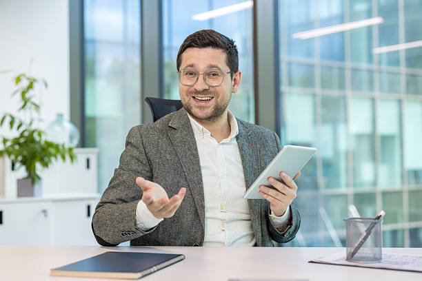 istock A businessman in a video conference, holding a tablet and gesturing in an office setting. He appears to be in a virtual meeting. 2225679660