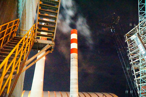 This image features an industrial landscape at night, showcasing a tall chimney emitting smoke. The scene includes intricate metal staircases, and construction machinery, highlighting the complexity