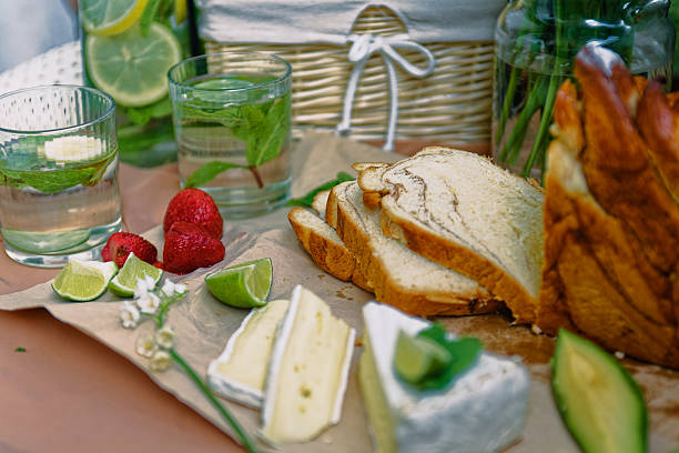 A Delicious Picnic Scene Featuring Freshly Baked Swirled Bread, Creamy Brie Cheese Garnished With Lime And Mint, Avocado Slices, Strawberries, Refreshing Citrus Drinks, And Blooming Lily Of The Valley Flowers Placed On A Rustic Paper Surface For A Natural Setting