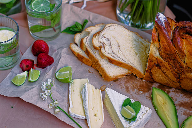 A Delicious Picnic Scene Featuring Freshly Baked Swirled Bread, Creamy Brie Cheese Garnished With Lime And Mint, Avocado Slices, Strawberries, Refreshing Citrus Drinks, And Blooming Lily Of The Valley Flowers Placed On A Rustic Paper Surface For A Natural Setting