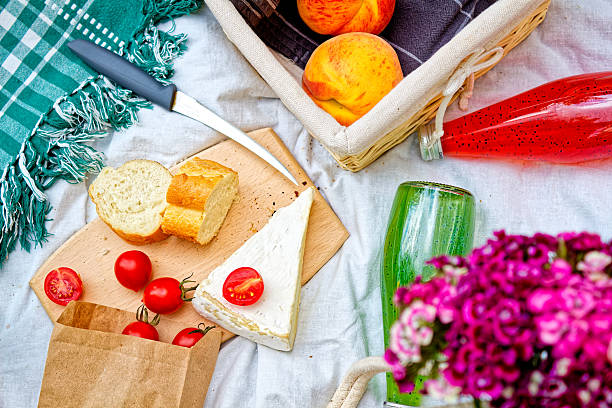 Summer Picnic Still Life With Baguette, Cheese, Tomato, Peach, Fresh Drink And Straw Hat On White Blanket. Romantic Outdoor Food Arrangement On Grass With Flowers, Natural Light And Rustic Textures