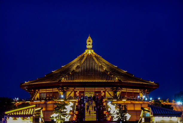 A sprawling, traditional-style hall in Luoyang, likely within Luoyi Ancient City, is beautifully lit at night. The golden illumination highlights the building's long, low profile and its series of elegant, curved roofs. The reflection on the wet ground in front adds depth and luster to the scene. This photo captures the immersive atmosphere of the reconstructed ancient city, making visitors feel as if they have stepped back in time into a prosperous ancient capital.