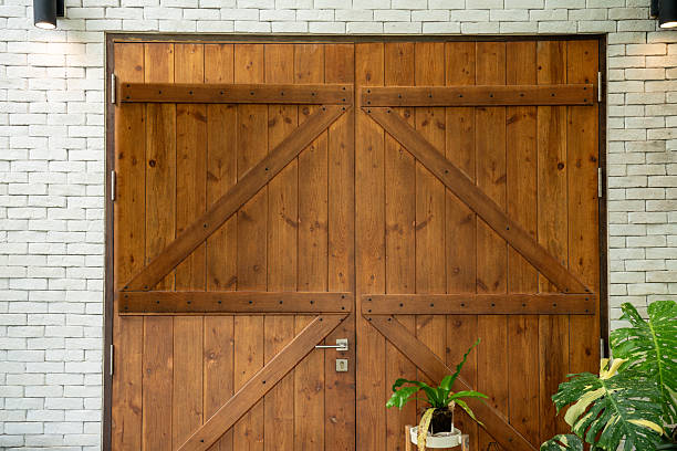 istock Rustic wooden double doors with diagonal cross braces, set in a white brick wall. Two modern black lights above and green potted plants below add a cozy, welcoming vibe. 2225705431