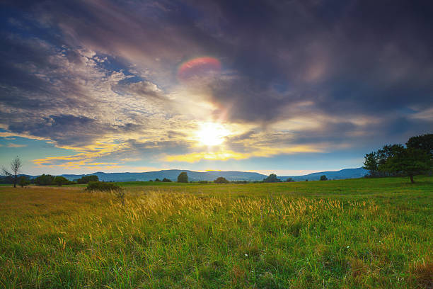 A vast grassy field stretches toward distant mountains under a dramatic sky filled with clouds, bright sunlight, and colorful sunset hues of gold and blue. This scene captures the raw beauty of changing weather patterns over agricultural land, symbolizing nature's constant cycle of storm and calm.