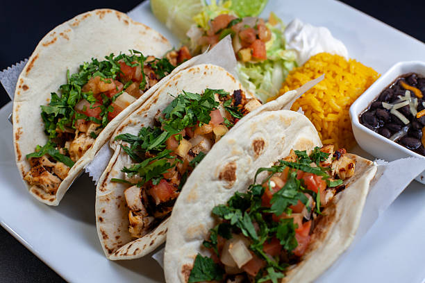 Close-up of a plate featuring three grilled chicken tacos topped with chopped cilantro, onions, and fresh salsa, served with a side of yellow rice, black beans, sour cream, lettuce, and lime wedges. A vibrant and appetizing presentation ideal for restaurant menus, food delivery apps, or Mexican cuisine content.