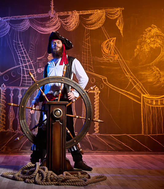 A cheerful black-bearded young man is dressed in a pirate costume. He is standing at the helm of a ship. Studio shot, the chalk drawing of a pirate ship floating in the sea on the blackboard as the background