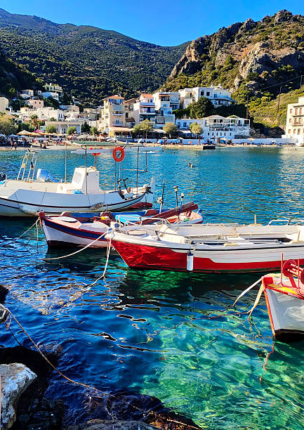 Idyllic spa village Therma, Ikaria island, Greece, with white houses along the seacoast, a bay with colorful boats and azure waters, picturesque surrounding hills, creating a serene atmosphere of Mediterranean culture and lifestyle
