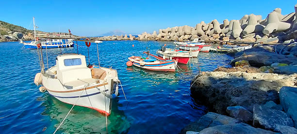 Idyllic spa village Therma, Ikaria island, Greece, with white houses along the seacoast, a bay with colorful boats and azure waters, picturesque surrounding hills, creating a serene atmosphere of Mediterranean culture and lifestyle