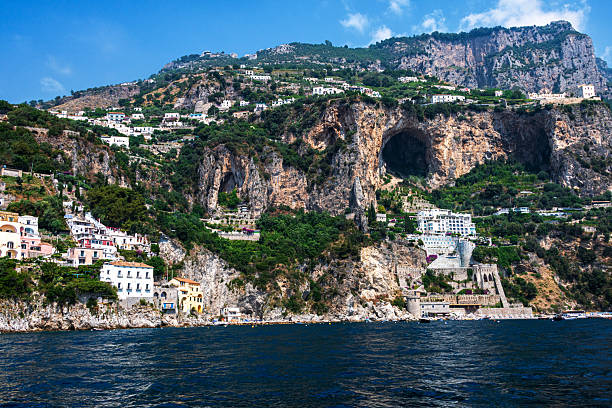A scenic coastal landscape of the Amalfi Coast, featuring rugged cliffs, turquoise waters, and Mediterranean vegetation. The image captures the natural beauty of the southern Italian coastline on a clear summer day, showcasing the region's iconic topography without focusing on any specific town or village.